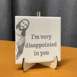 May include: White ceramic tile with a black and white image of a person's face and the text "I'm very disappointed in you". The tile is on a wooden stand.