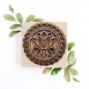A layered wooden lotus mandala with light and dark wood tones rests on a square piece of light brown paper, surrounded by green leaves.