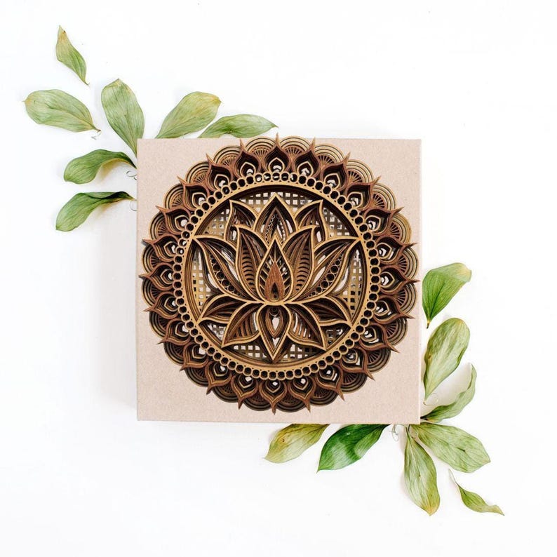 A layered wooden lotus mandala with light and dark wood tones rests on a square piece of light brown paper, surrounded by green leaves.
