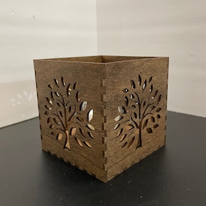 Peut inclure: Un bougeoir en bois brun foncé avec un motif d'arbre découpé sur chaque côté. Le bougeoir est de forme carrée et a une finition lisse.