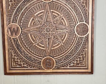 May include: Wooden wall art featuring a layered compass design. The compass rose is centered within a square frame, with the cardinal directions labeled N, W, E, and S. The artwork is crafted from light and dark wood tones.