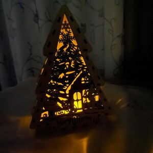 May include: A wooden Christmas tree cutout with a house and snowflakes inside. The tree is lit from within, casting a warm glow. The words "Merry Christmas" are visible at the bottom of the tree.