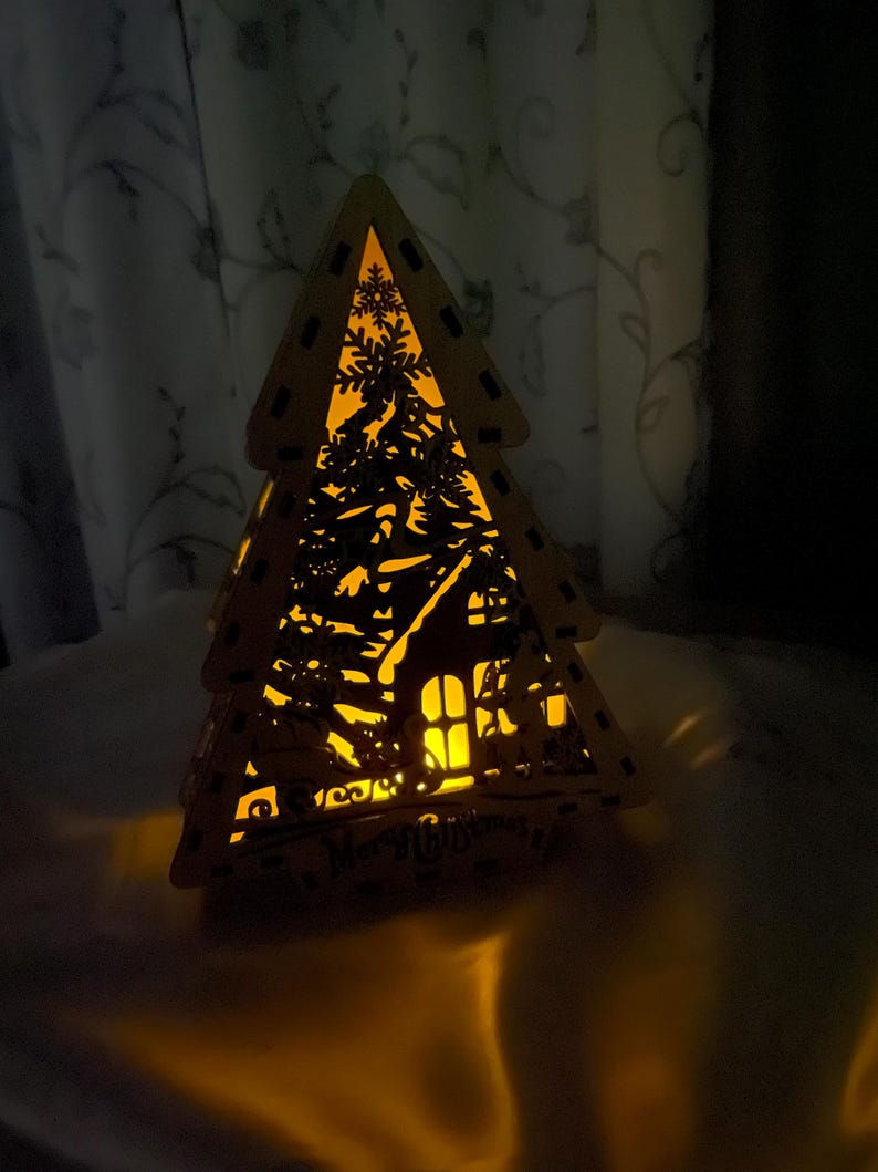 May include: A wooden Christmas tree cutout with a house and snowflakes inside. The tree is lit from within, casting a warm glow. The words "Merry Christmas" are visible at the bottom of the tree.