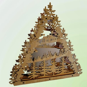 A laser-cut wooden Christmas decoration, shaped like a Christmas tree, displays a layered winter scene with deer, trees, and a house, viewed from an angle.