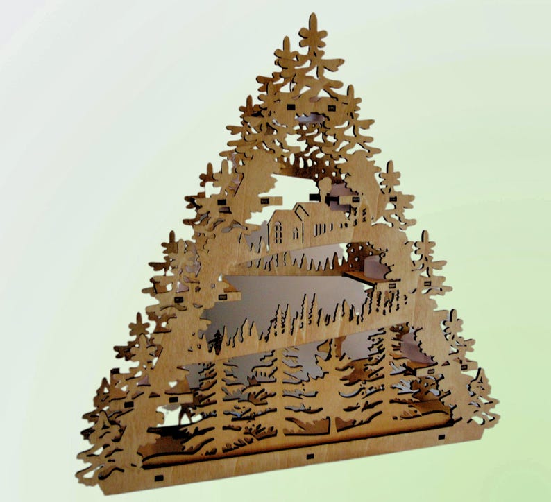 A laser-cut wooden Christmas decoration, shaped like a Christmas tree, displays a layered winter scene with deer, trees, and a house, viewed from an angle.