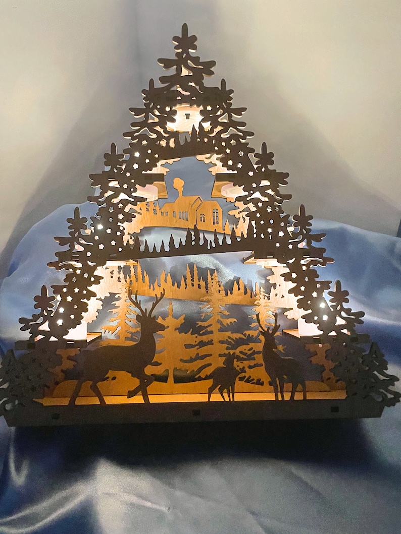 K&ouml;nnte beinhalten: Eine Weihnachtsdorf-Szene aus Holz mit einem beleuchteten Haus, B&auml;umen und Rehen. Die Szene ist aus lasergeschnittenem Holz gefertigt und strahlt ein warmes, einladendes Licht aus.