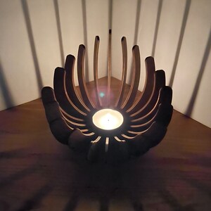 K&ouml;nnte beinhalten: Ein Kerzenhalter aus Holz in Form einer Blume mit einer brennenden Kerze in der Mitte. Der Halter steht in einer Ecke und wirft Schatten auf die Holzoberfl&auml;che.