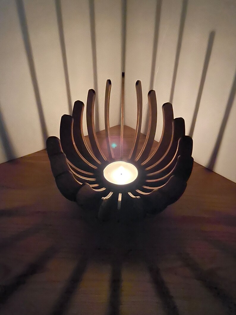 K&ouml;nnte beinhalten: Ein Kerzenhalter aus Holz in Form einer Blume mit einer brennenden Kerze in der Mitte. Der Halter steht in einer Ecke und wirft Schatten auf die Holzoberfl&auml;che.