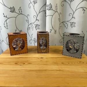 Three wooden tealight holders, each with a tree of life cutout on two sides, in light brown, medium brown, and gray, sit on a light wood table.