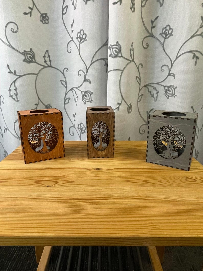 Three wooden tealight holders, each with a tree of life cutout on two sides, in light brown, medium brown, and gray, sit on a light wood table.