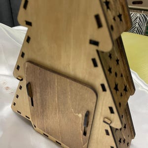 May include: A wooden Christmas tree decoration with a star-shaped cutout design. The ornament is stained brown and has a small, rectangular opening on the back.