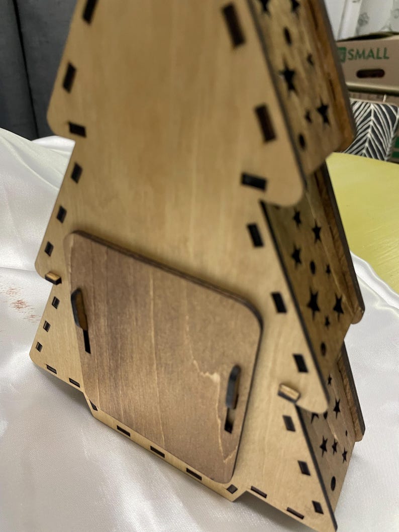 May include: A wooden Christmas tree decoration with a star-shaped cutout design. The ornament is stained brown and has a small, rectangular opening on the back.
