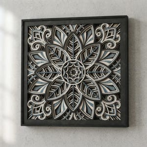 May include: A square wall art piece featuring a multi-layered mandala design with intricate leaf-like petals and swirling patterns. The artwork is composed of stacked layers in shades of charcoal gray, light gray, and white, enclosed in a black frame.