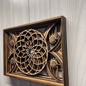 Puede incluir: Una pieza de arte mural de madera en capas con un dise&ntilde;o geom&eacute;trico. La obra de arte tiene un marco marr&oacute;n oscuro y capas intrincadas de color marr&oacute;n claro y beige, creando un efecto 3D. El dise&ntilde;o central es un patr&oacute;n circular con acentos florales.