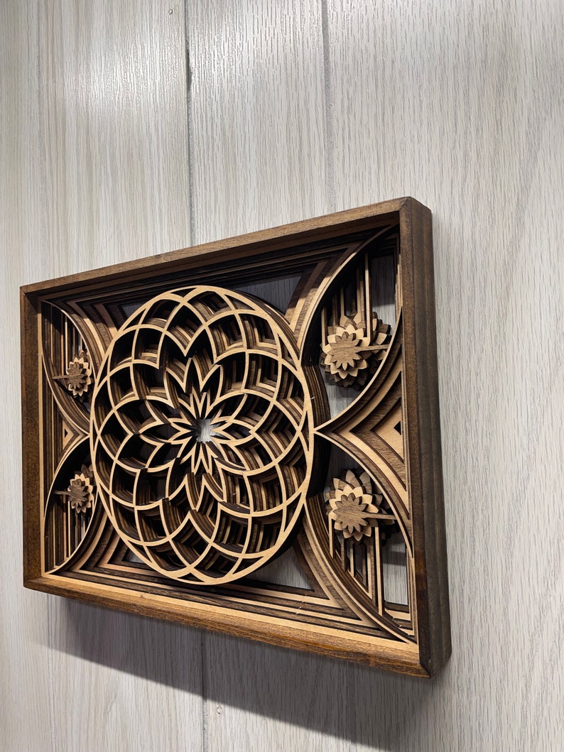 Puede incluir: Una pieza de arte mural de madera en capas con un dise&ntilde;o geom&eacute;trico. La obra de arte tiene un marco marr&oacute;n oscuro y capas intrincadas de color marr&oacute;n claro y beige, creando un efecto 3D. El dise&ntilde;o central es un patr&oacute;n circular con acentos florales.