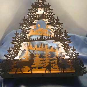 A laser-cut wooden Christmas decoration with a layered winter scene of deer, trees, and a house is brightly lit from behind by seven flameless tea lights.