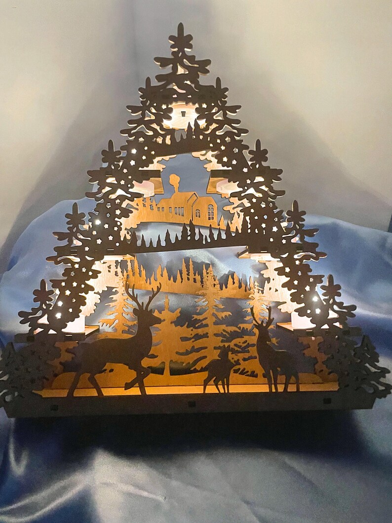 A laser-cut wooden Christmas decoration with a layered winter scene of deer, trees, and a house is brightly lit from behind by seven flameless tea lights.