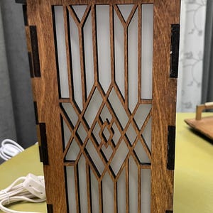 Puede incluir: Una lámpara de madera con un diseño geométrico y un panel de vidrio esmerilado blanco. La lámpara tiene un acabado marrón oscuro y un cable negro.