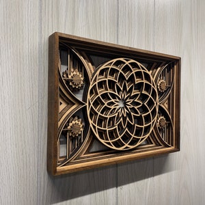 Puede incluir: Una pieza de arte mural de madera en capas con un dise&ntilde;o floral geom&eacute;trico. La obra de arte est&aacute; construida a partir de m&uacute;ltiples capas de madera, creando un efecto tridimensional. El marco es de color marr&oacute;n oscuro y las capas interiores son de un tono de madera m&aacute;s claro.