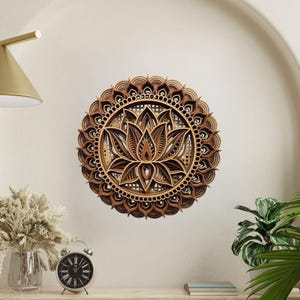 A layered wooden lotus mandala with light and dark wood tones hangs on a light beige wall above a wooden shelf with a clock and plants.