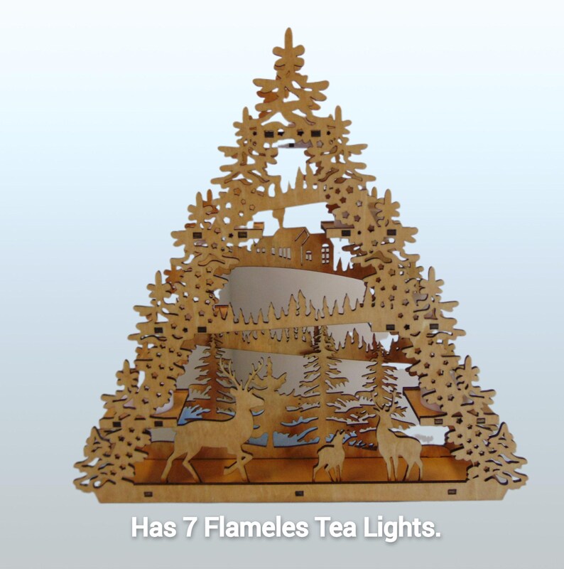A laser-cut wooden Christmas decoration, shaped like a tree, shows a layered winter scene with deer, trees, and a house, with seven flameless tea lights.