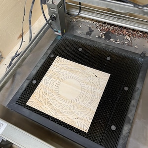 A laser cutter bed holds a single intricately cut light-colored wooden mandala square.