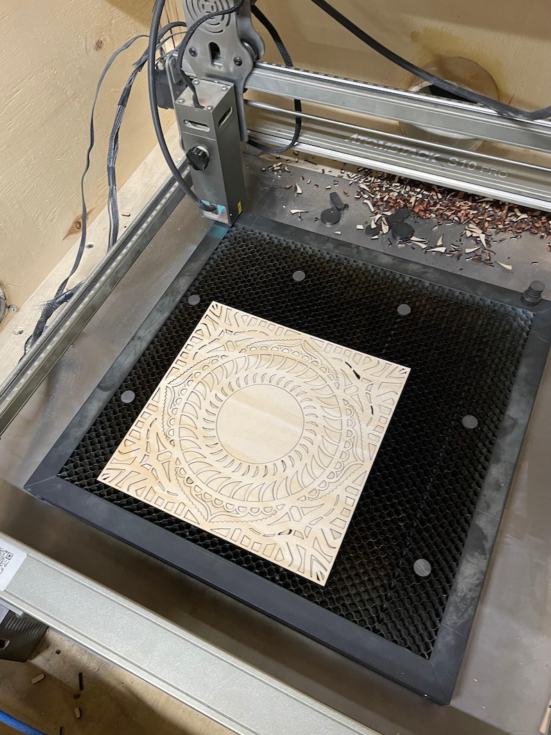 A laser cutter bed holds a single intricately cut light-colored wooden mandala square.