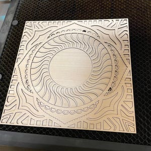 Close-up view of an intricately laser-cut light wood square panel featuring circular mandala and geometric patterns.