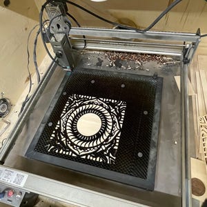 A laser cutting machine bed holds a black-finished intricately cut square mandala layer.