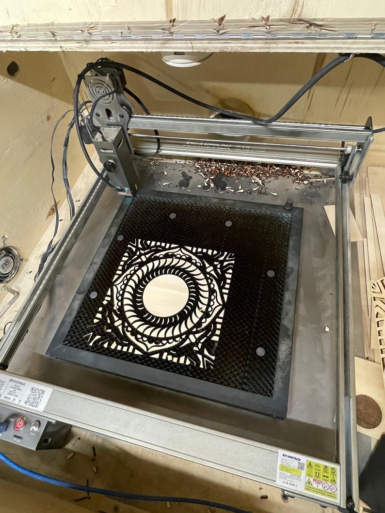 A laser cutting machine bed holds a black-finished intricately cut square mandala layer.
