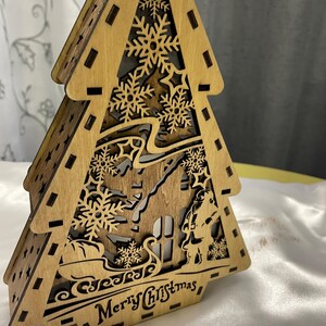 May include: A wooden Christmas tree cutout with a house, a figure, and snowflakes. The tree is lit up and says "Merry Christmas" at the bottom.