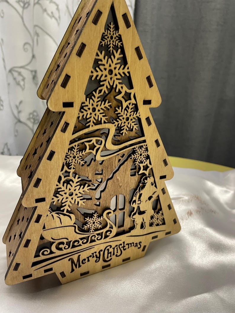 May include: A wooden Christmas tree cutout with a house, a figure, and snowflakes. The tree is lit up and says "Merry Christmas" at the bottom.