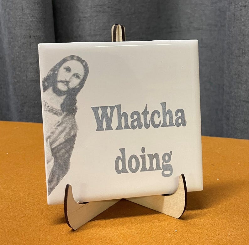 A white ceramic tile with a black and white image of Jesus peeking from the left side, and the words "Whatcha doing" in gray text, displayed on a light wood stand.
