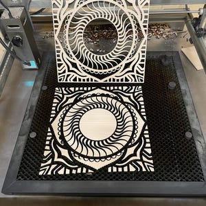 A laser engraving machine bed holds a light-colored cut wooden mandala layer, with a matching cut piece held upright above it.