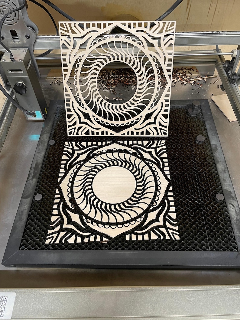A laser engraving machine bed holds a light-colored cut wooden mandala layer, with a matching cut piece held upright above it.