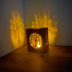 A medium brown wooden tealight holder with a tree of life cutout casts a warm, tree-shaped shadow on a dark wall and table.