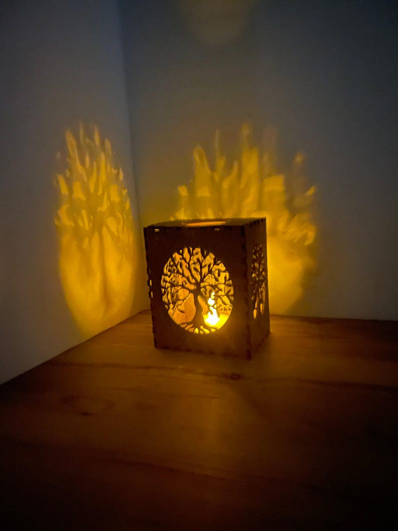 A medium brown wooden tealight holder with a tree of life cutout casts a warm, tree-shaped shadow on a dark wall and table.