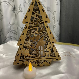 May include: A wooden Christmas tree cutout featuring a Santa Claus and a house design. The tree is illuminated by a small white LED tea light.