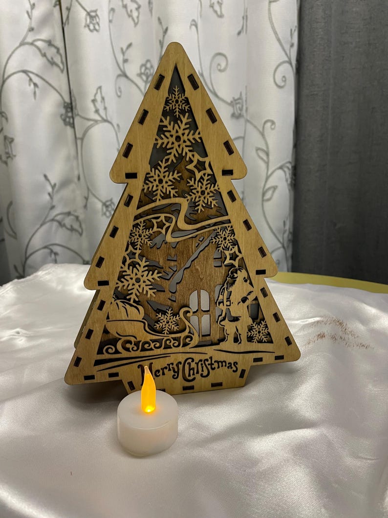 May include: A wooden Christmas tree cutout featuring a Santa Claus and a house design. The tree is illuminated by a small white LED tea light.
