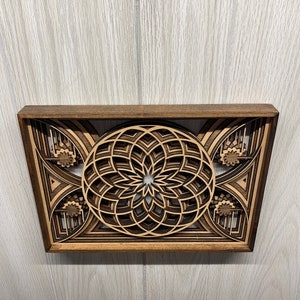 Puede incluir: Una pieza de arte mural de madera en capas con un dise&ntilde;o floral central y patrones geom&eacute;tricos. La obra de arte est&aacute; enmarcada en madera marr&oacute;n oscuro, con tonos de madera m&aacute;s claros que crean profundidad y contraste. El dise&ntilde;o es intrincado y detallado.