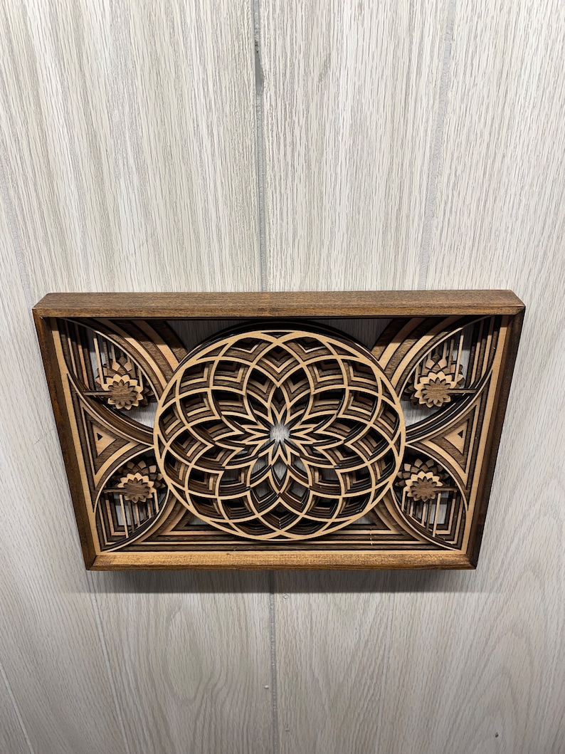 Puede incluir: Una pieza de arte mural de madera en capas con un dise&ntilde;o floral central y patrones geom&eacute;tricos. La obra de arte est&aacute; enmarcada en madera marr&oacute;n oscuro, con tonos de madera m&aacute;s claros que crean profundidad y contraste. El dise&ntilde;o es intrincado y detallado.