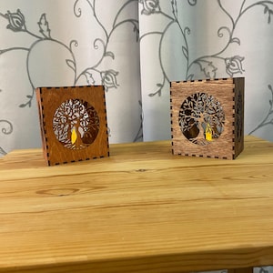 Two wooden tealight holders, each with a tree of life cutout on two sides, in light brown and medium brown, sit on a light wood table.