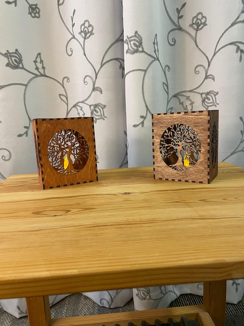Two wooden tealight holders, each with a tree of life cutout on two sides, in light brown and medium brown, sit on a light wood table.