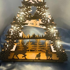 A laser-cut wooden Christmas decoration with a layered winter scene of deer, trees, and a house is illuminated from behind by seven flameless tea lights.