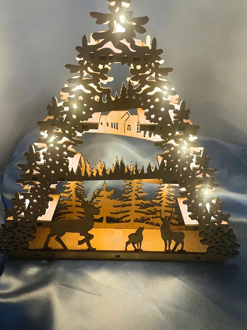 A laser-cut wooden Christmas decoration with a layered winter scene of deer, trees, and a house is illuminated from behind by seven flameless tea lights.