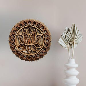 A layered wooden lotus mandala with light and dark wood tones hangs on a light beige wall next to a white vase holding dried palm leaves.