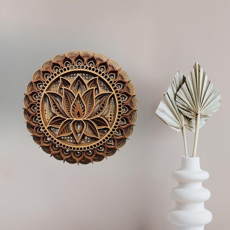 A layered wooden lotus mandala with light and dark wood tones hangs on a light beige wall next to a white vase holding dried palm leaves.