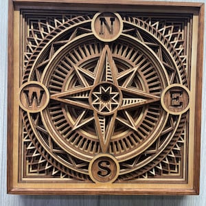 May include: A square, multi-layered wooden wall art piece featuring a central compass rose design. The artwork displays intricate geometric patterns in varying shades of brown wood, with the letters N, S, E, and W positioned at the cardinal points.