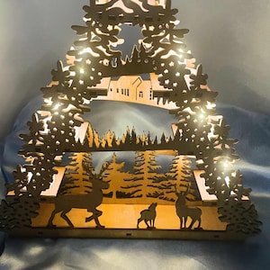A laser-cut wooden Christmas decoration with a layered winter scene of deer, trees, and a house is brightly illuminated by seven flameless tea lights, casting a warm glow.