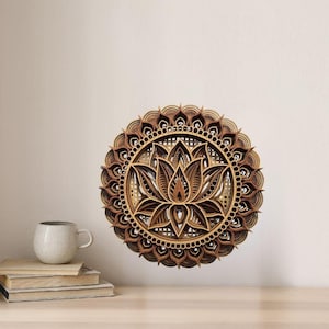 A layered wooden lotus mandala with light and dark wood tones hangs on a light beige wall above a wooden table with a white mug and books.